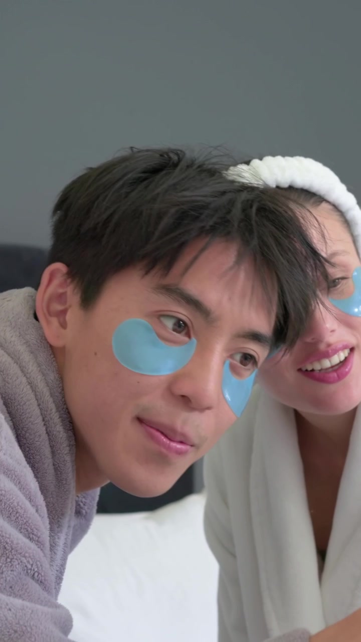 Vertical video: Leaning couple checking mirror on bed, adjusting blue under-eye patches in robes