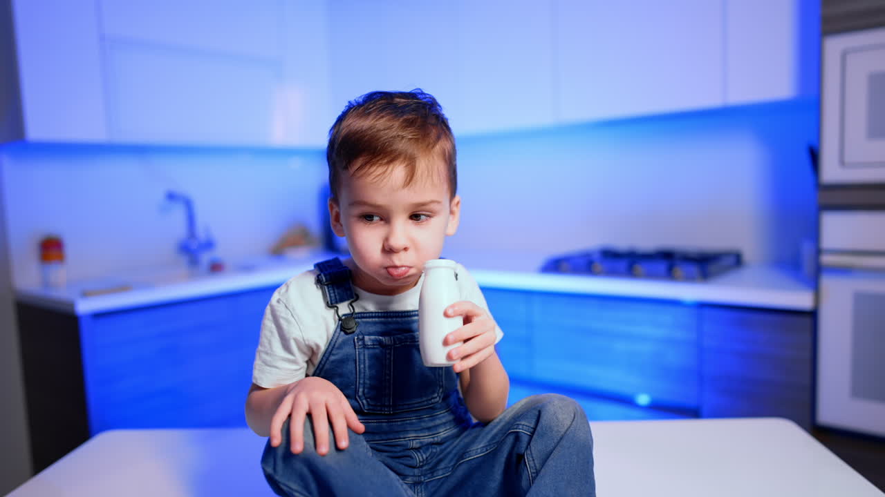 Little kid in white t-shirt and jeans romper sits on the kitchen table. Cute baby boy drinks dairy from a plastic bottle.