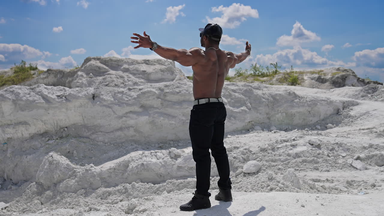Muscular dark skinned man without shirt stands on white hill. Shirtless african athlete stands with stretched arms among summer nature. Sportsman in the mountains. Rear view.