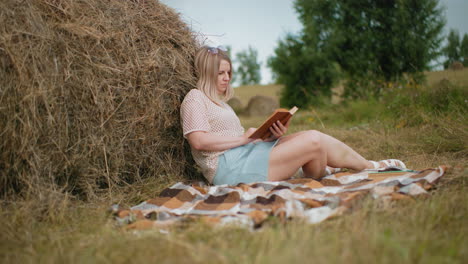 side view of thoughtful woman seated on blanket, reading book near hay bale in open farmland, green trees, rolling hills, and a cloudy sky surround her