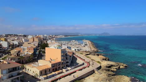 experimente la serena belleza de la playa de son baulo en can picafort, mallorca, con aguas azules claras, costas rocosas y vibrantes edificios costeros bajo un cielo soleado.