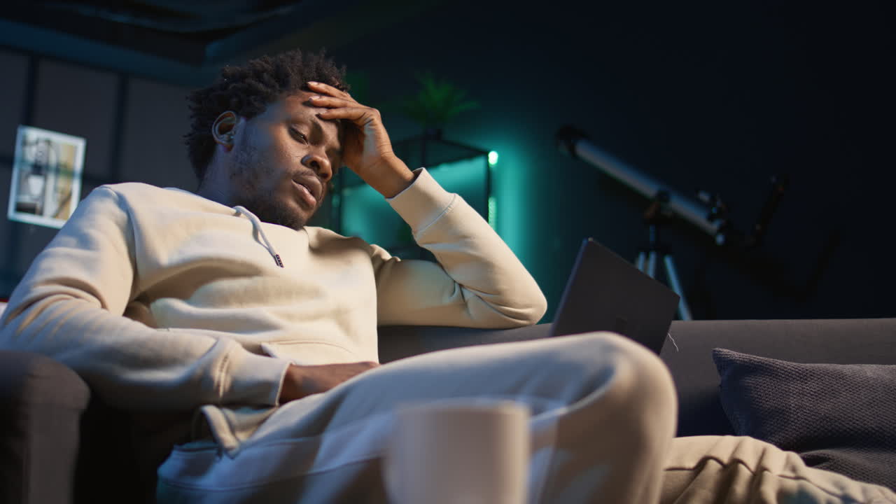 Self employed man suffering from pain and high fever sitting on sofa, trying to work and finish his tasks. Black male feelancer suffering from illness and feeling sick, using laptop. Camera A.