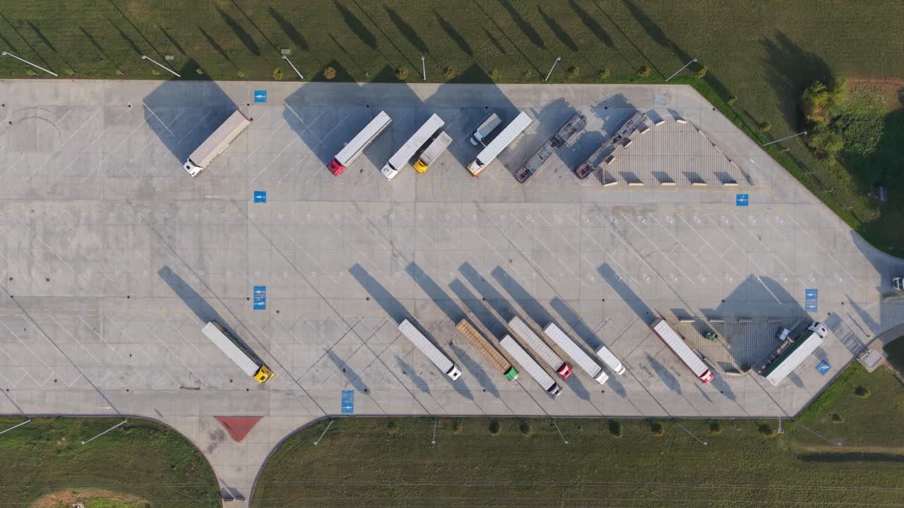 A top-down aerial view of a truck parking lot showing multiple semi-trailers parked in designated spaces. The image captures the geometric layout of the lot and the long shadows cast by the vehicles