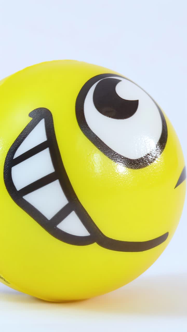 Una palla di gomma gialla con faccina sorridente rotola su uno sfondo bianco sotto una luminosa illuminazione da studio