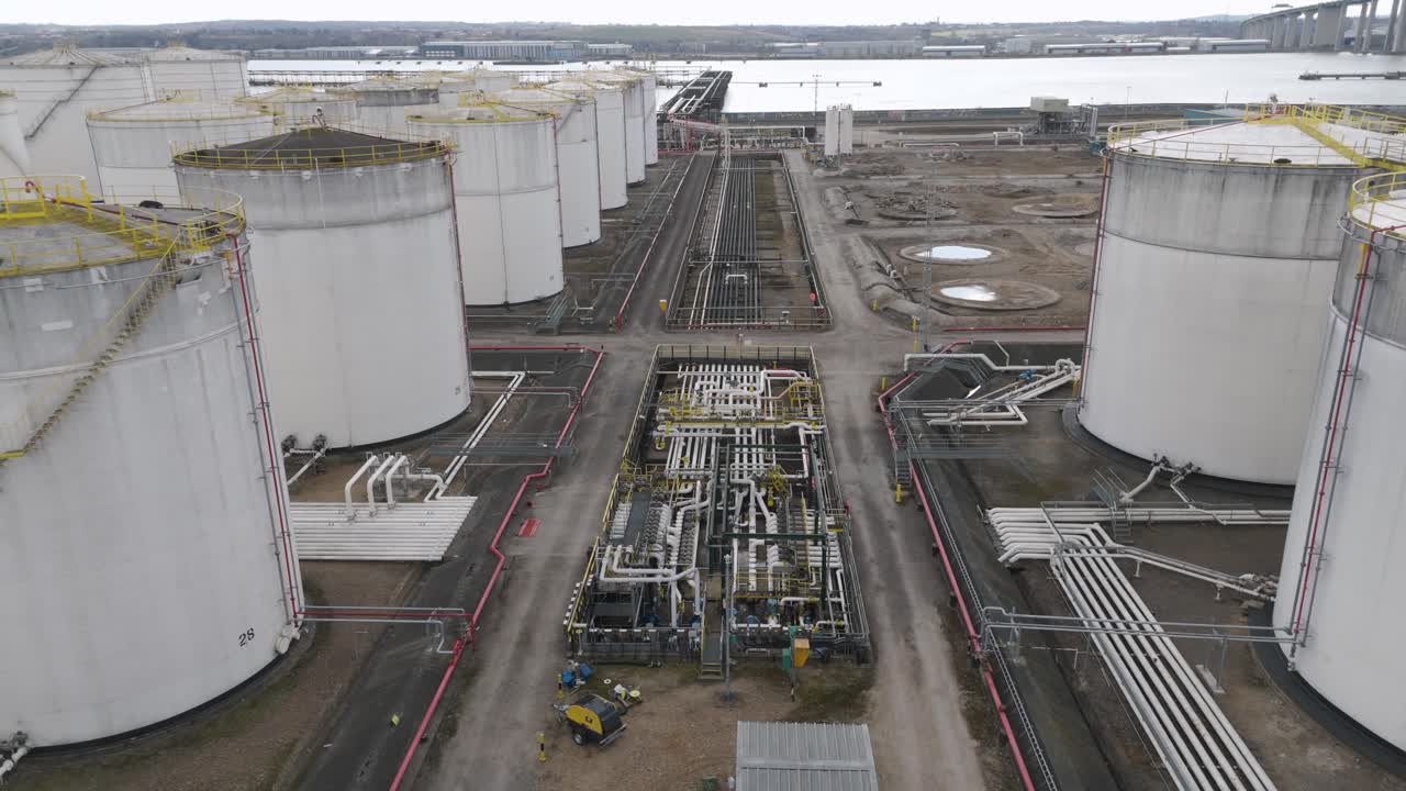 Elevated aerial of riverside gas holders lined in a row with flat domes, service roads and pipelines illustrating energy-storage and distribution network