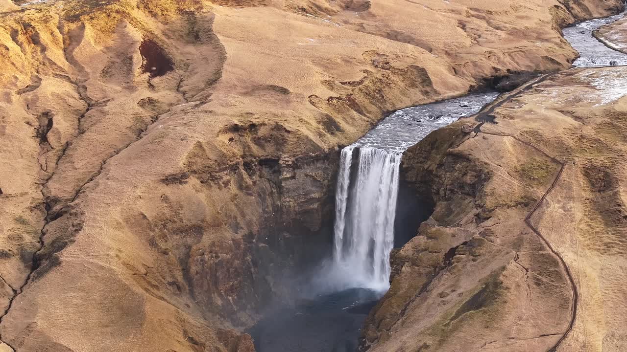 Aerial view of Skógafoss waterfall in Iceland, where the Skógá river dramatically drops from the ancient sea cliffs into a deep gorge. Barren golden hills near Skógar and the Eyjafjallajökull volcano
