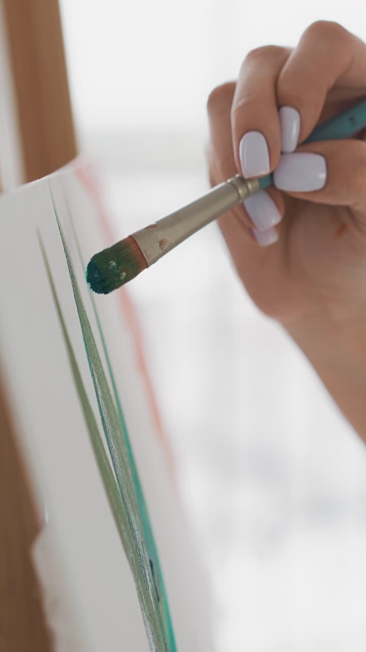 Elegant lady hand draws lush tropical leaf with small paintbrush on white canvas fixed on wooden easel in light art studio extreme closeup