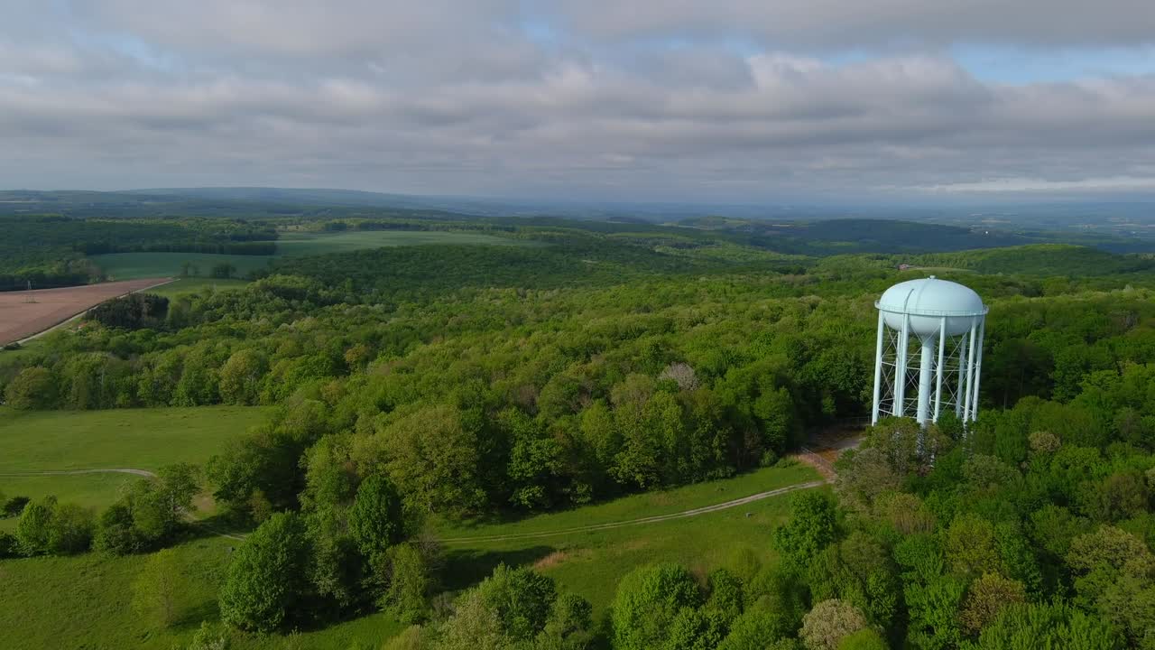 Aerial drone view of white water tower rising above dense forest on Chestnut Ridge near Morgantown, surrounded by rolling hills and greenery under partly cloudy sky