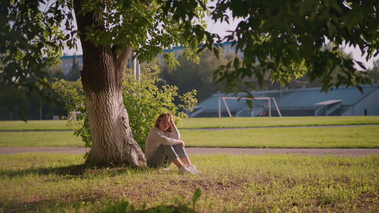 나무 아래 잔디에 사려 깊게 앉아 있는 여자, 머리와 다리에 손을 고 상하는 자세로, 배경에는 초록색 잔디, 기둥, 경기장 등이 있다
