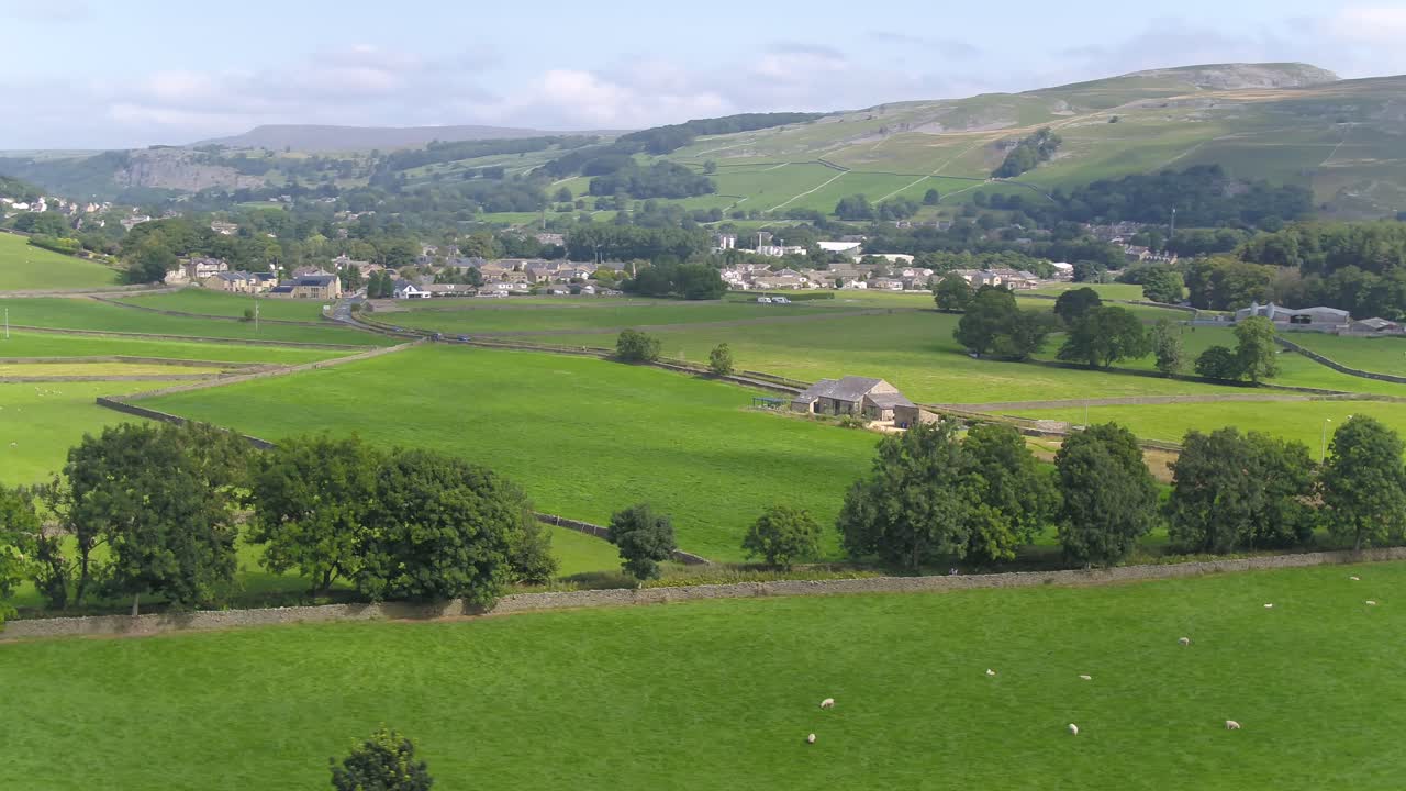 Drone footage moving sideways and panning over farmland and fields near the rural countryside village of Settle, Yorkshire, UK with sheep, dry stone walls, trees with houses and a hilly landscape