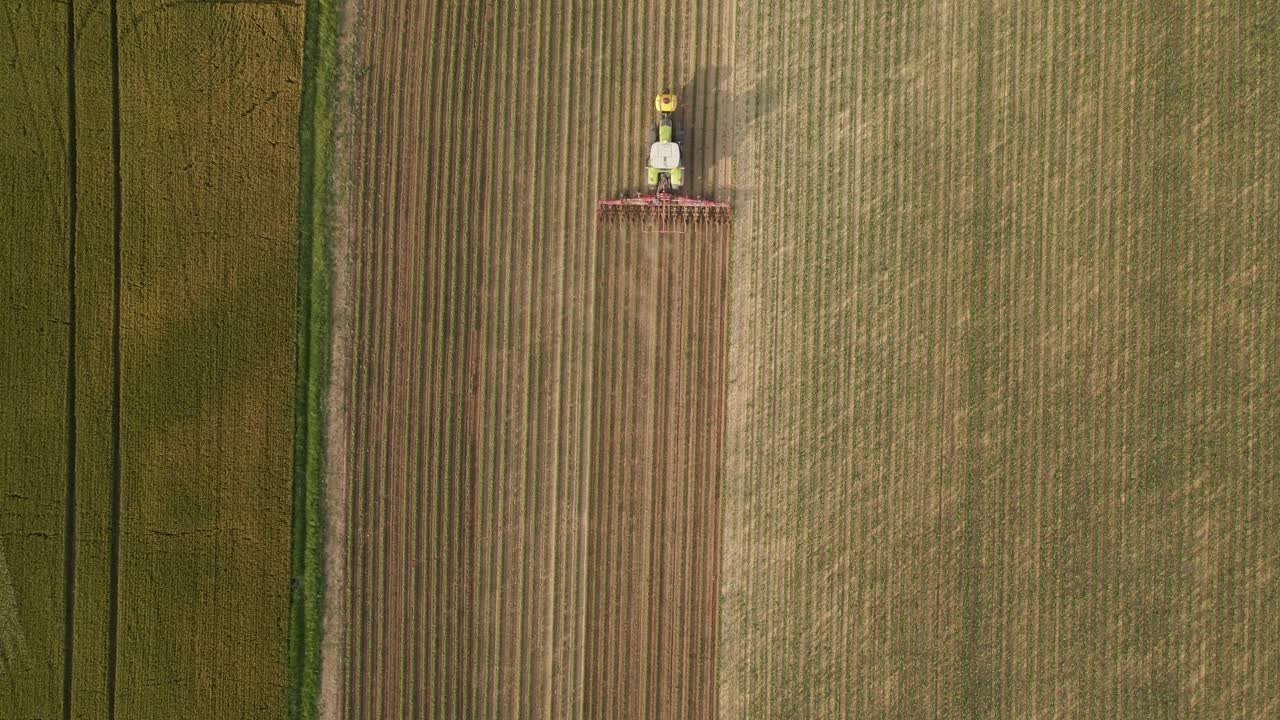 Aerial top-down view of a tractor cultivating a field with precise, evenly spaced rows. The machinery creates a striking striped pattern across the agricultural landscape.
