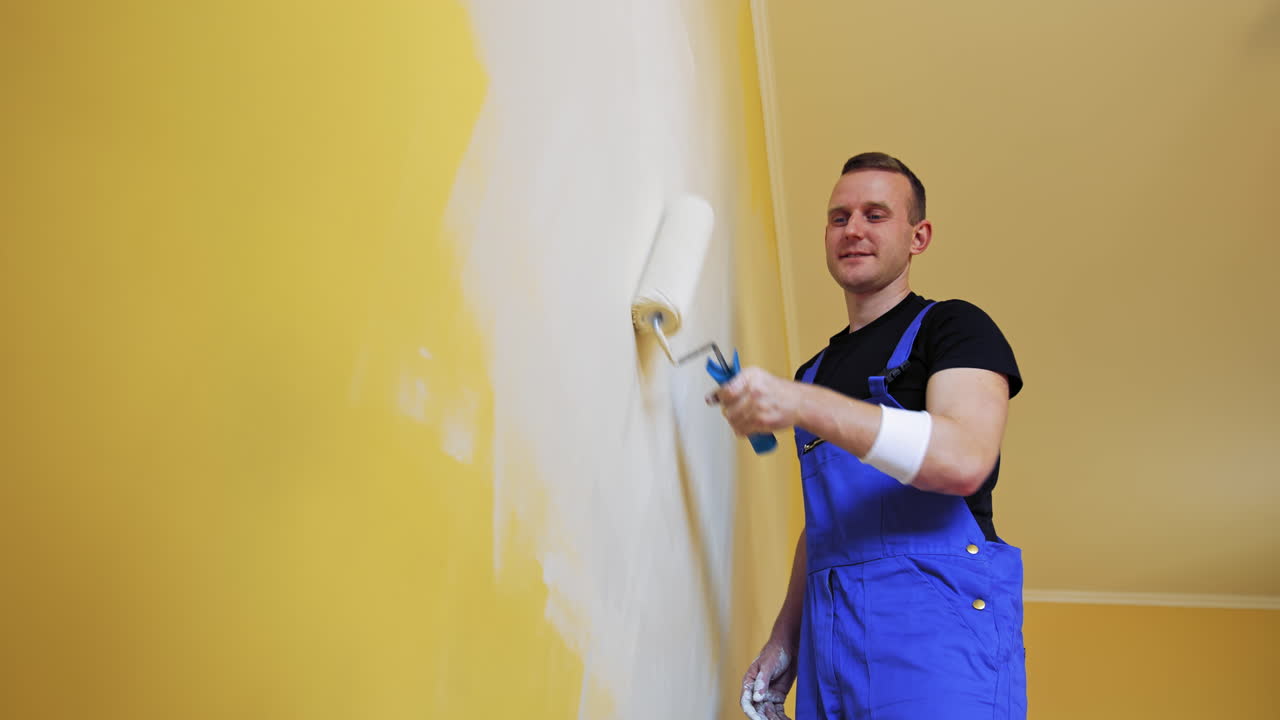 Roller repainting wall into light color. Young man in blue overalls painting wall into white in the apartment. Makeover in flat.