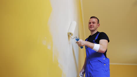 roller repainting wall into light color. young man in blue overalls painting wall into white in the apartment. makeover in flat.