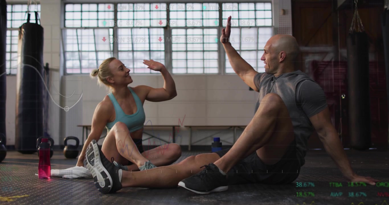 Training duo sitting on gym floor, talking building, high-fiving with data charts celebrating gains