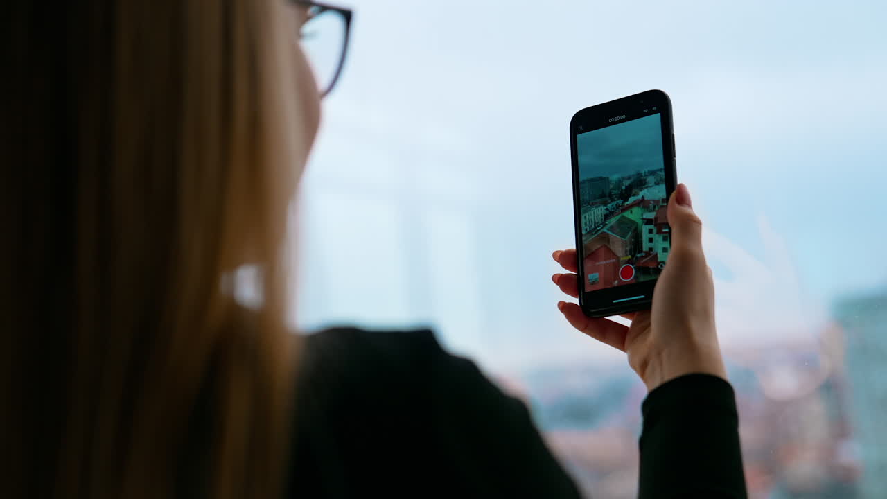 Girl with a mobile phone indoors. Young woman taking photo of a cityscape through the office window. Close-up.