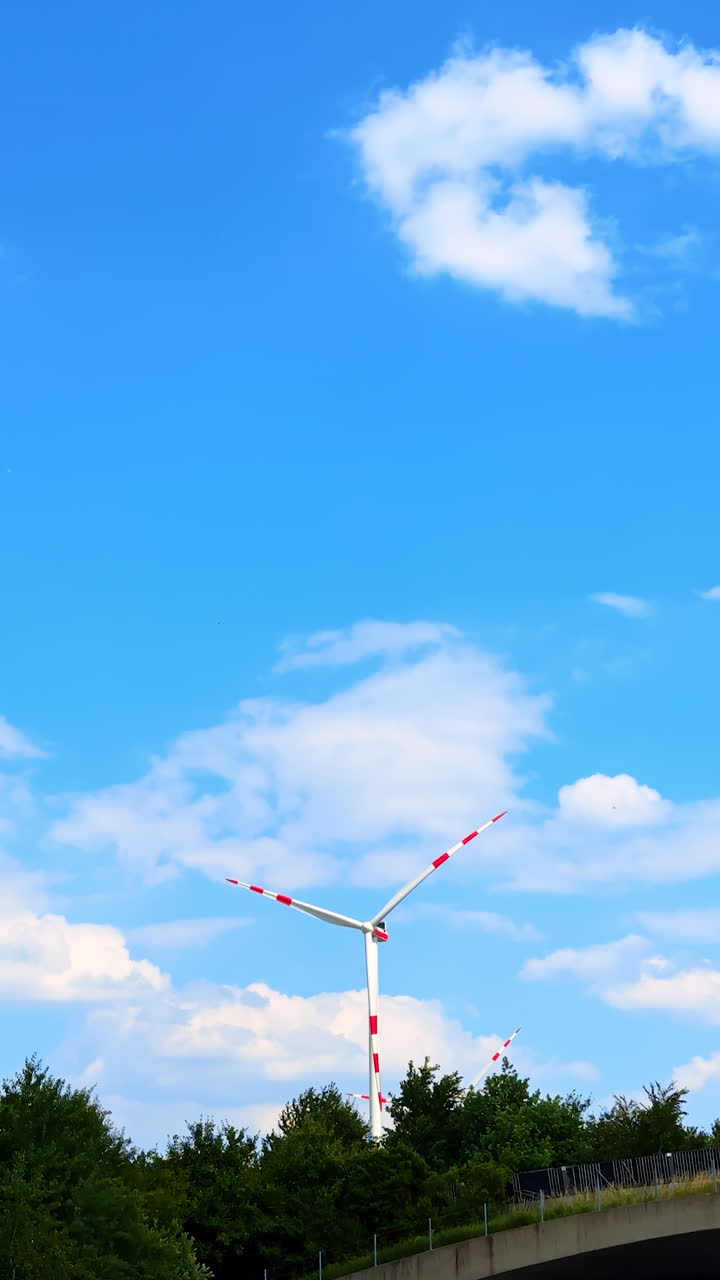 Wind turbine spinning under blue sky. A wind turbine spins gently against a blue sky with clouds, framed by lush greenery