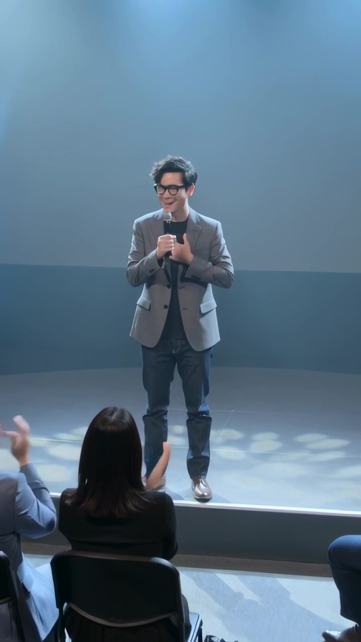 Video verticale: Uomo asiatico con blazer grigio che parla, tiene un microfono e si inchina mentre il pubblico applaude sul palco.