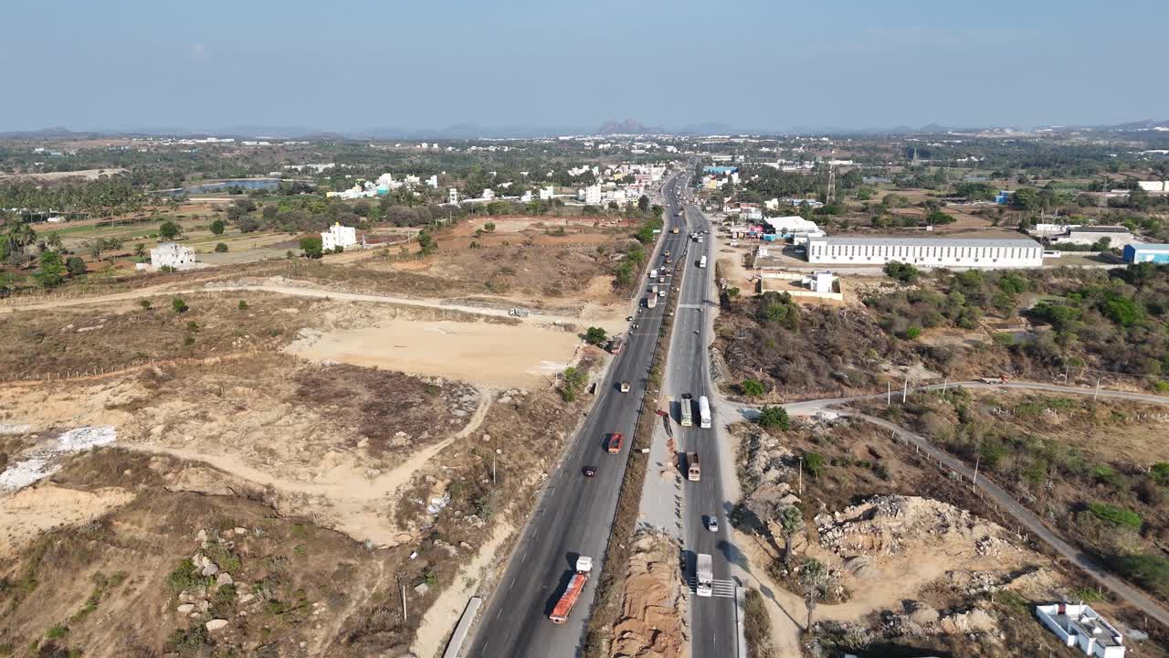 Aerial view of a highway under construction next to an active factory, warehouse and a small town. Traffic flows, with rolling hills in the background under a clear sky