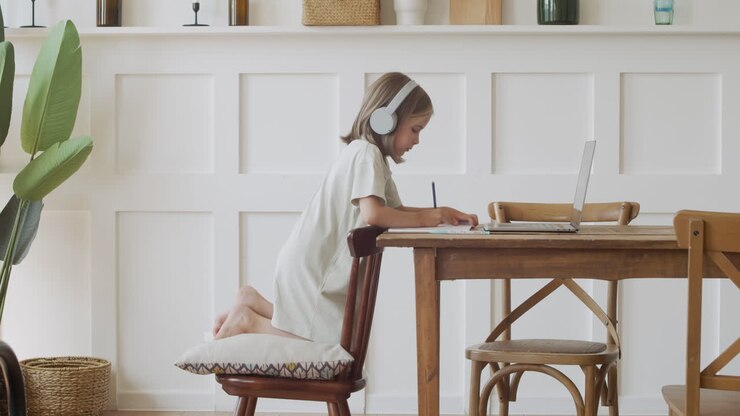 A Responsible Cute Blonde Girl With Headphones Looks At The Laptop Screen And Does Her Homework Using A Pencil And Paper