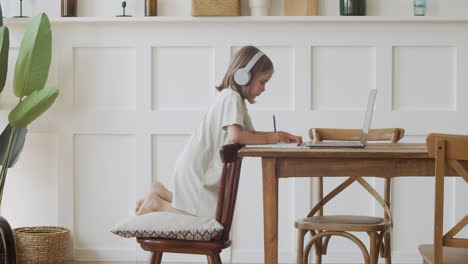 A-Responsible-Cute-Blonde-Girl-With-Headphones-Looks-At-The-Laptop-Screen-And-Does-Her-Homework-Using-A-Pencil-And-Paper