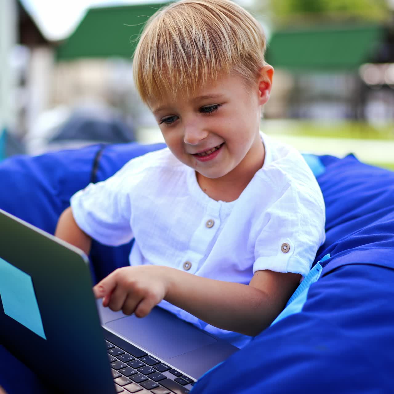 Caucasian six-year old with laptop outdoors. Peaceful boy in bean bag chair outdoors playing computer. Blurred backdrop