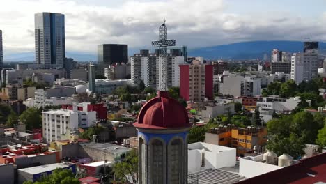 vertigo effect of church tower, steeple, red dome, and steel cross in downtown mexico city center buildings, skyscrapers and skyline with mountain range in background on cloudy day, aerial zoom in