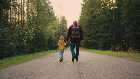 little boy and his grandfather are walking together on road in forest in spring or autumn day rear view