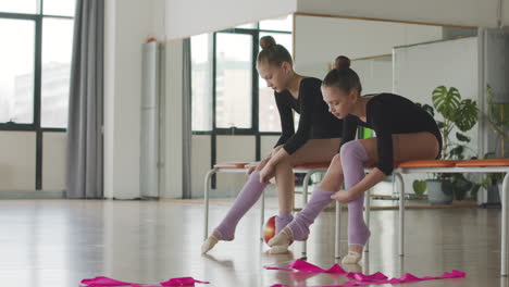 Zwei-Gymnastische-Blonde-Mädchen,-Die-Ihre-Socken-Anziehen,-Sitzen-Auf-Einer-Bank,-Bevor-Sie-Mit-Der-Ballettklasse-2-Beginnen