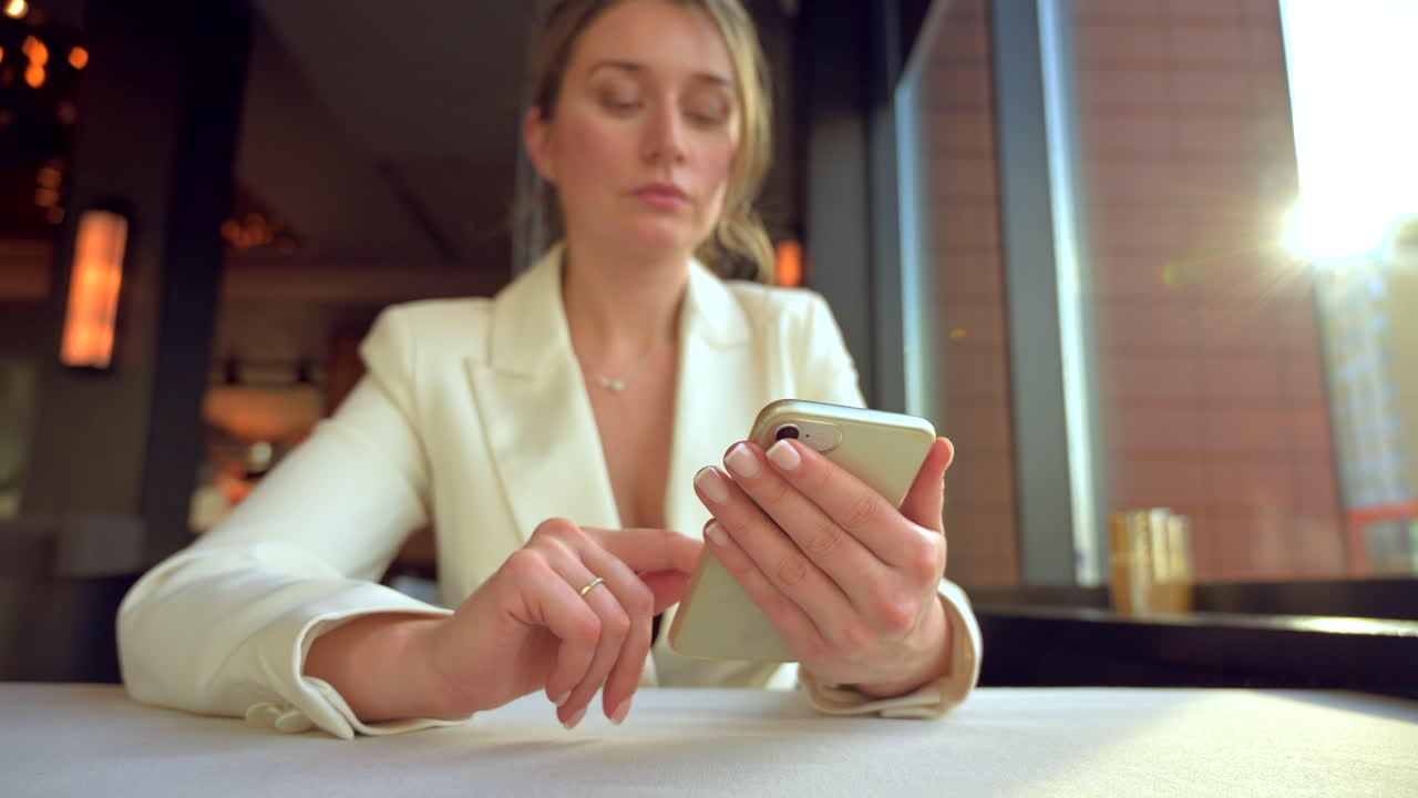 Woman in a white blazer scrolling on her phone at a table at a restaurant