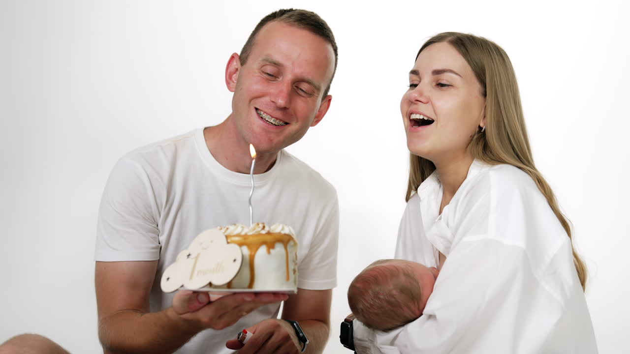 Smiling Caucasian couple with baby blowing a candle on the cake. Family celebrating first month of their infant. White backdrop.