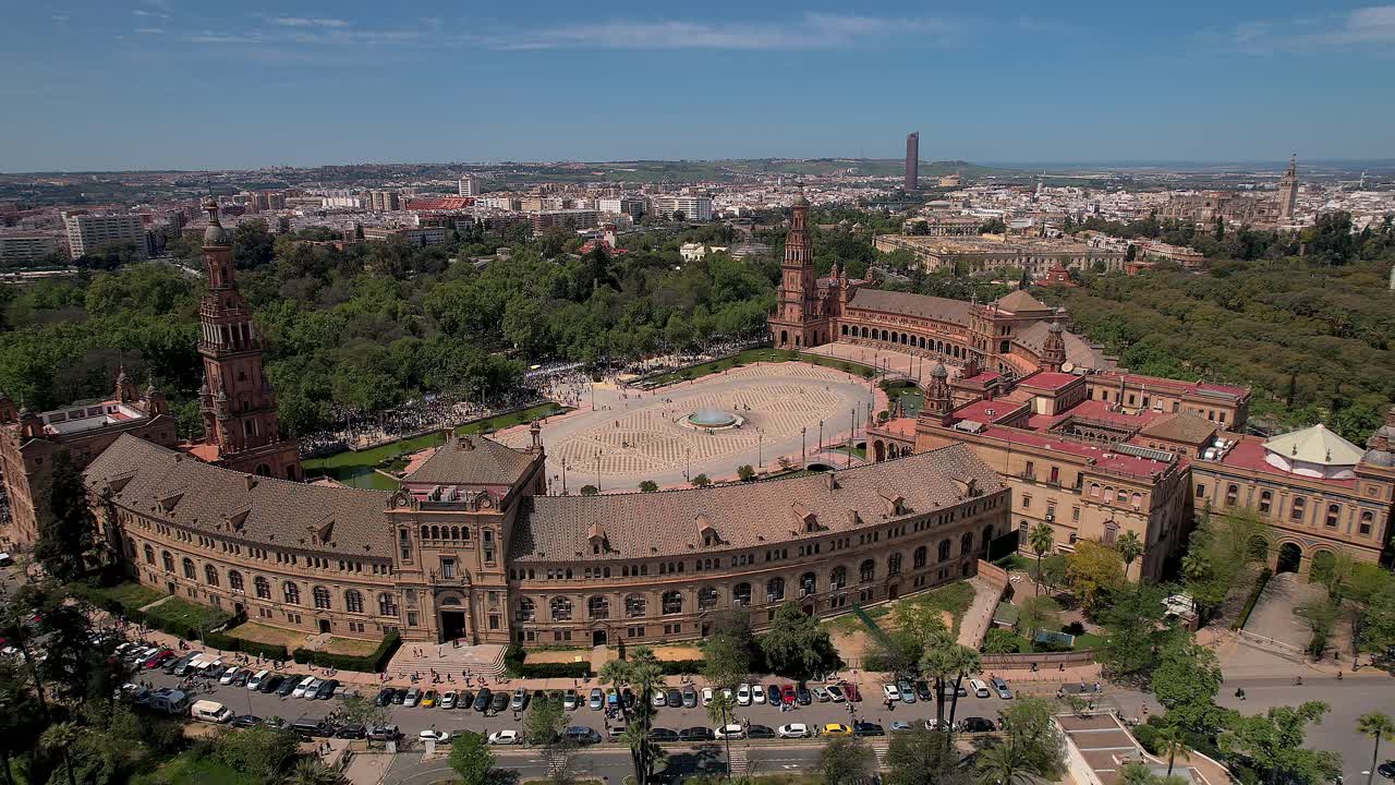 Wide establishing aerial shot of Plaza de España in Parque de María Luisa, Seville, Spain, showing the semi-circular Renaissance Revival complex, central fountain and square with Torre Sevilla visible