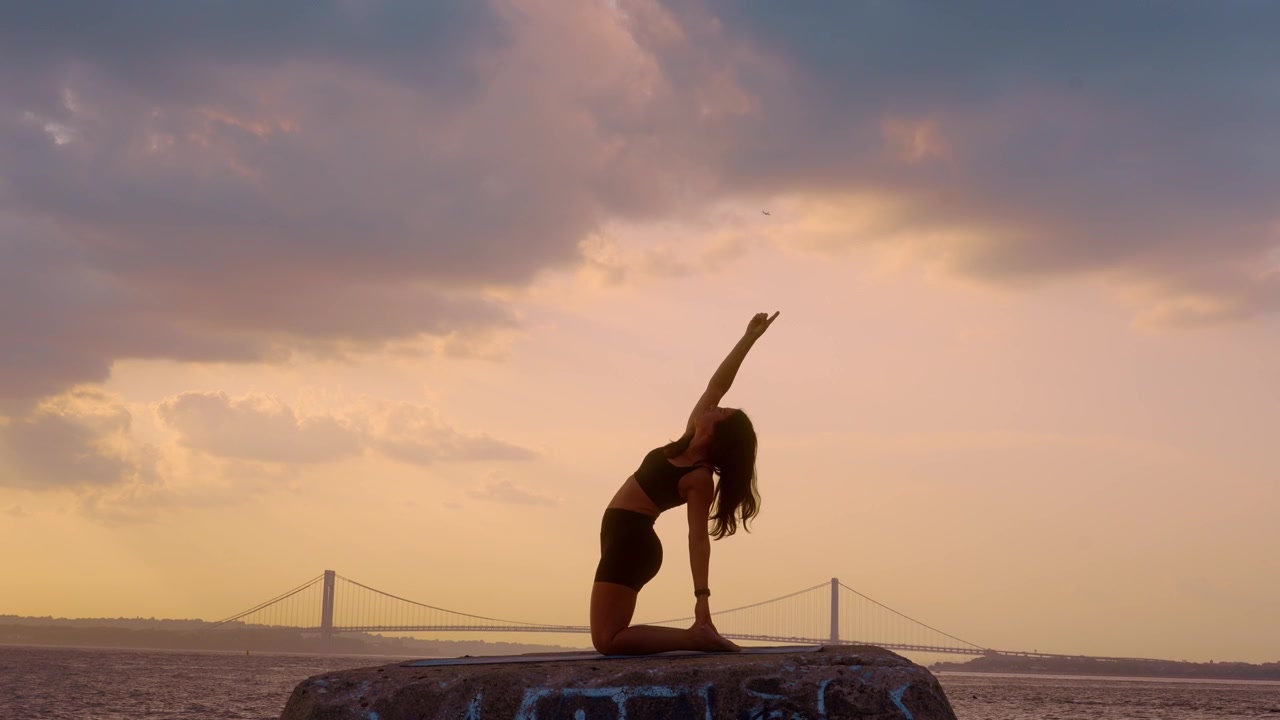 Donna che esegue un esercizio yoga di piegamento all'indietro in ginocchio al tramonto a New York. Profilo laterale con le braccia incrociate su un blocco lungomare, sullo sfondo il cielo del Ponte di Verrazzano Narrows.