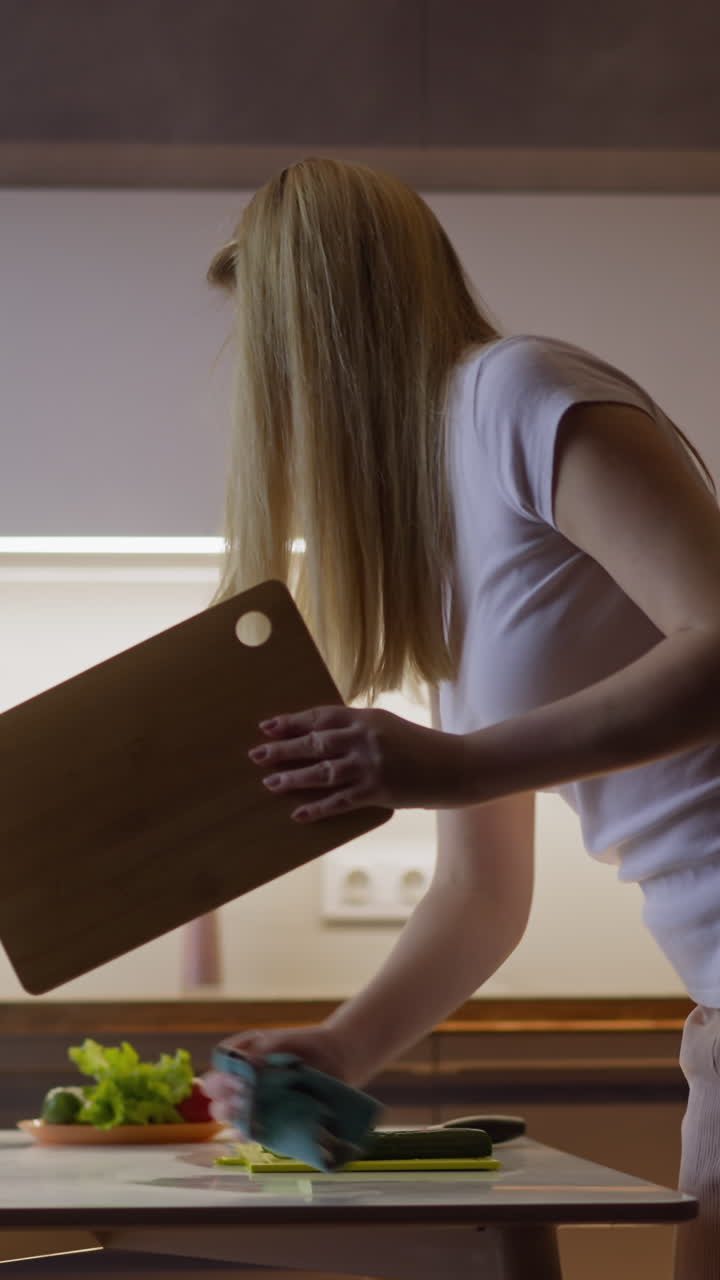 Long haired blonde woman in casual clothes cleans table with rug before beginning to cook dinner for family in modern kitchen side view slow motion