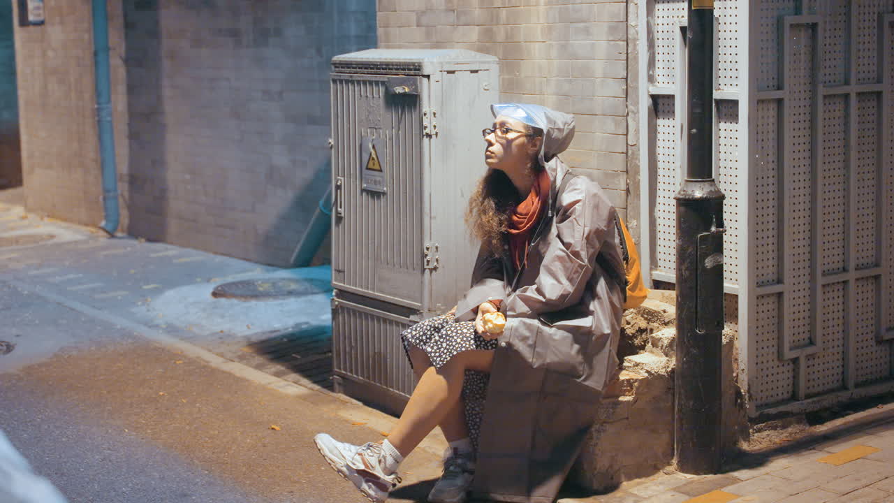 Asian woman sitting on sidewalk under lamp, tired commuter resting against utility box in hooded coat, hands clasped, vintage film tones, melancholic nighttime glow and wet asphalt texture