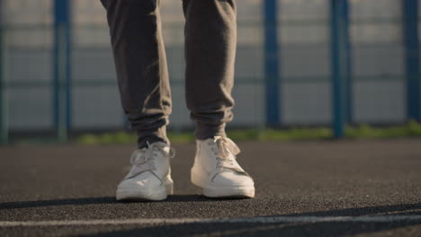 vista de las piernas de personas con ropa deportiva y zapatos de lona saltando y moviéndose lateralmente en la cancha con la luz del sol reflejándose en ellos, el fondo presenta un edificio e instalación deportiva