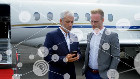 animación de una red de iconos digitales sobre dos hombres de negocios caucásicos usando un teléfono inteligente en el aeropuerto.
