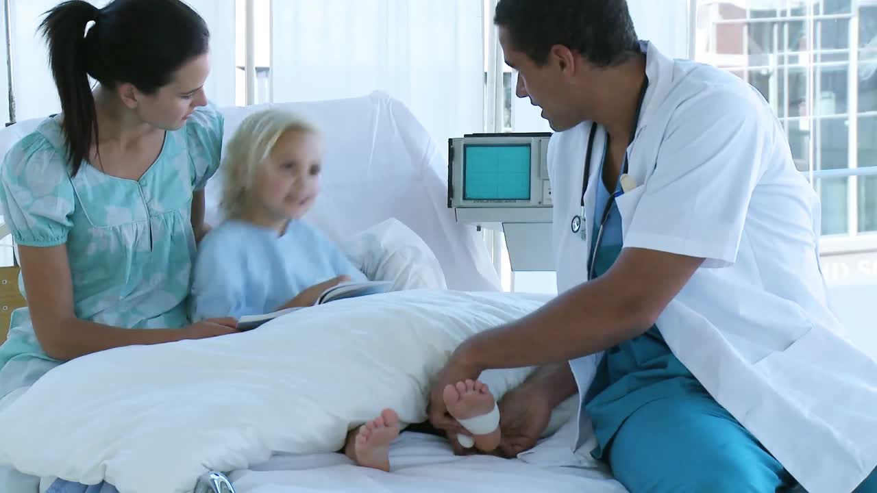 Doctor bandaging a little patients foot lying in bed with her mother