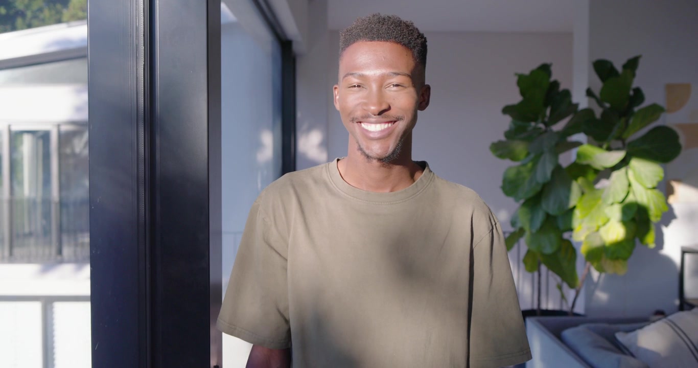 Afroamerikansk mand står nær glasdør, solen fanger blikket, vender sig mod kameraet, smilende og grinende.