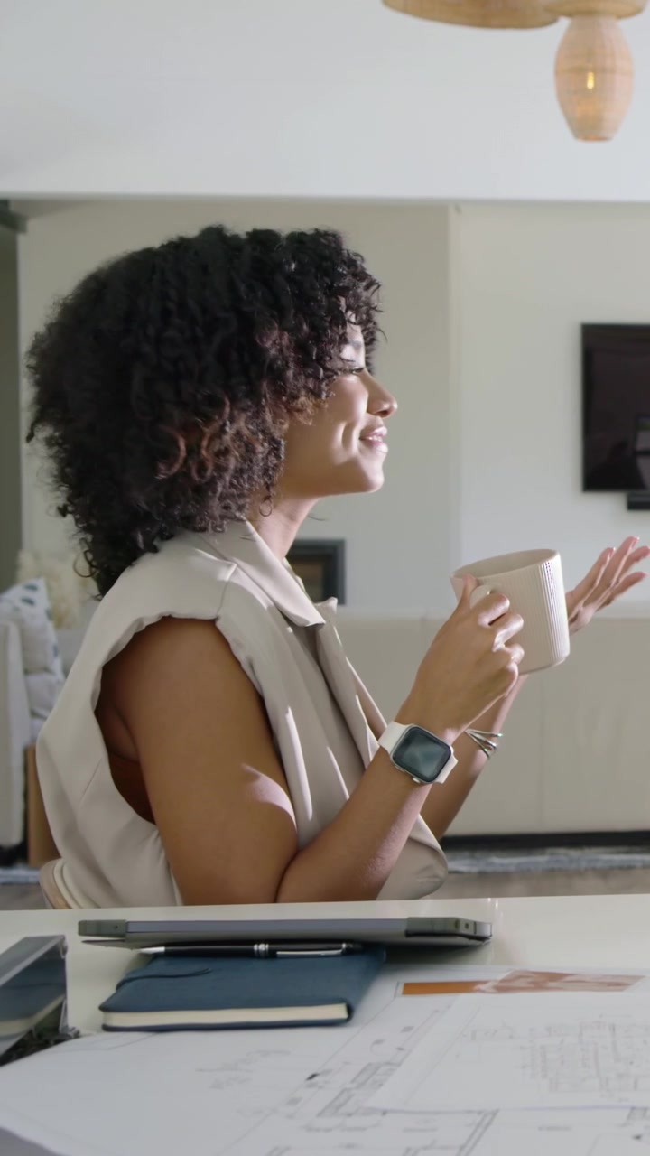 Vertical video: Smiling woman in beige vest sipping mug during virtual call at desk, laptop nearby