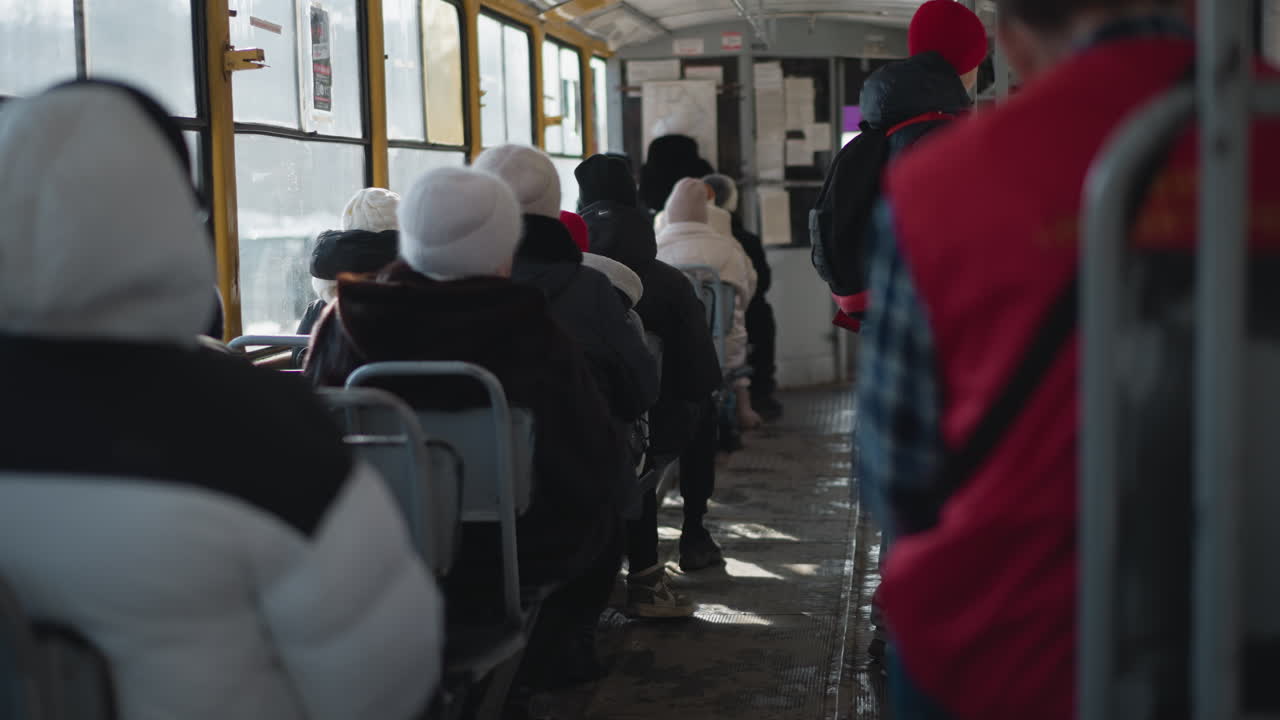 Interior de tren abarrotado con viajeros de invierno sentados mientras un pasajero está de pie agarrado a un poste mientras el vagón frena hacia la parada, luz fría a través de ventanas y marcos metálicos creando un ambiente de tránsito urbano crudo.