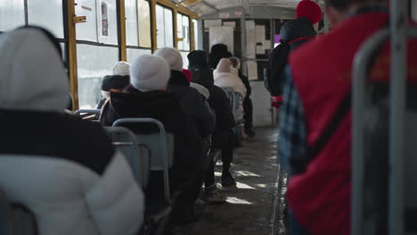 crowded train interior with winter commuters seated while one rider stands gripping pole as carriage slows toward stop, cold light through windows and metal frames creating gritty urban transit mood