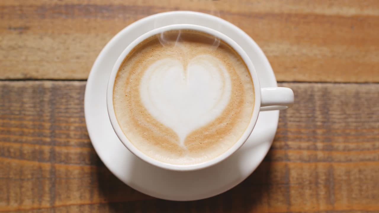 Tazza bianca fumante su piattino che rilascia vapore, mentre un latte si raffredda su un tavolo di legno con schiuma a forma di cuore.