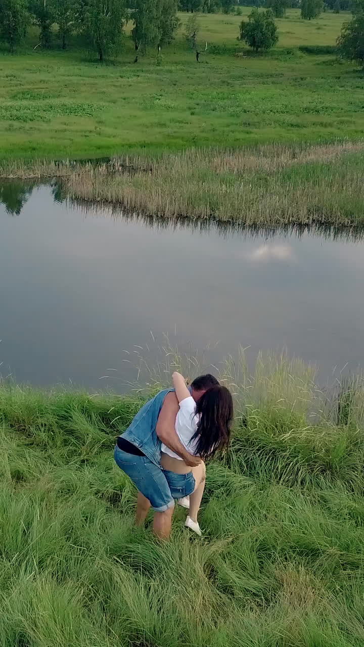 upper view man in denim vest and shorts spins brunette girl holding in arms on green meadow near blue lake