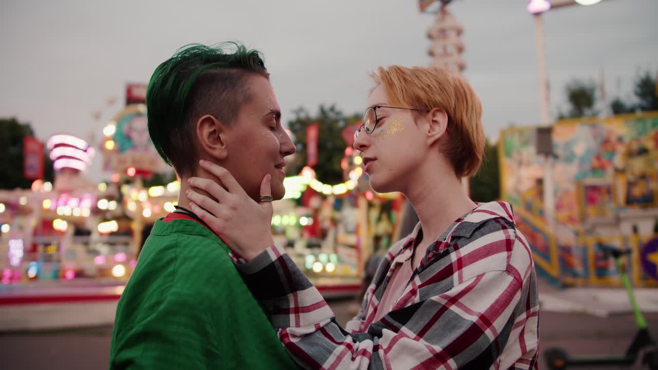 A blonde girl with a short hair strokes her girlfriend's head with green hair and a green shirt with both hands and then puts her head on her shoulder during her date in the amusement park