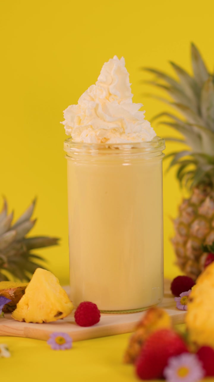Una mano aggiunge panna montata a un frullato all'ananas con frutta fresca, sfondo giallo brillante, inquadratura statica
