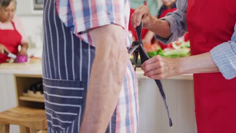 Happy-caucasian-senior-couple-putting-on-aprons,-in-kitchen-with-diverse-friends,-slow-motion