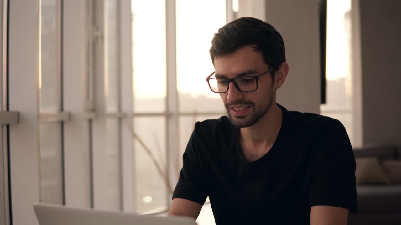 retrato de un hombre alegre usando una computadora portátil en un lugar de trabajo remoto en cámara lenta. autónomo trabajando en la computadora en casa. hombre de buen aspecto escribiendo activamente en la computadora portátil, hablando con amigos en una casa grande con ventanas panorámicas en el fondo