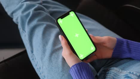 young woman sitting on sofa at home and uses smartphone with green screen mock-up. holding in hand and looking on the screen. 4k