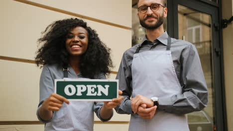 Portrait of smiling caucasian waiter and african american waitress standing holding signboard open" outside of coffee shop"