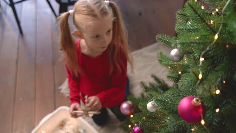 vista superior de la niña rubia arrodillada en el suelo recogiendo la decoración navideña de una caja y colgándola en el árbol de navidad